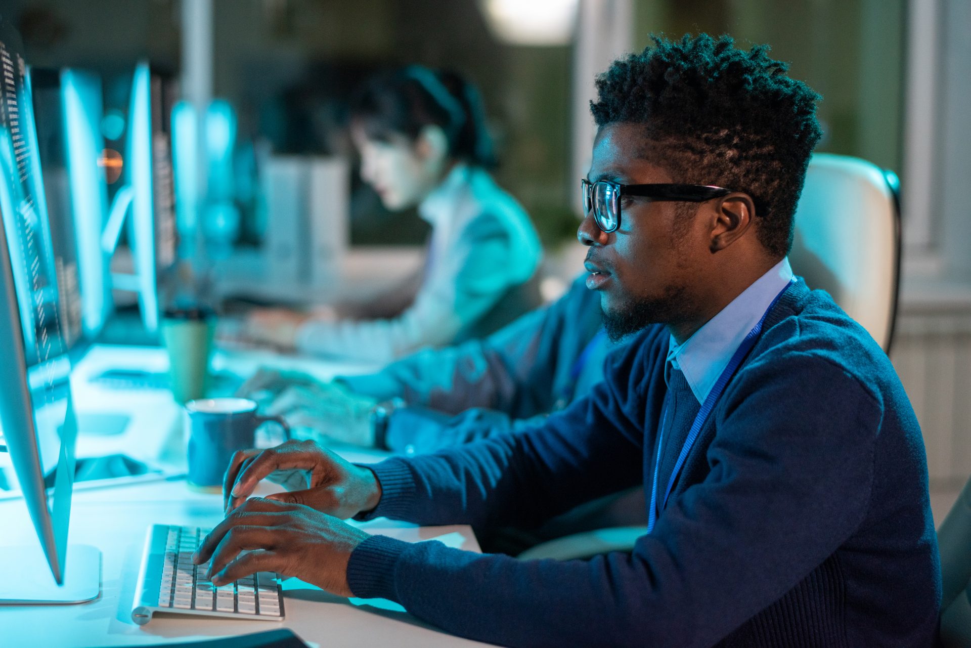 Young serious businessman in eyeglasses and formalwear looking at screen of computer against his coworkers