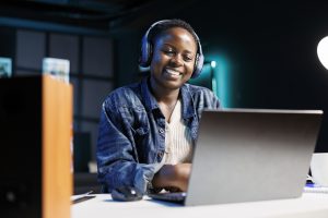 Cheerful black woman uses wireless headphones while browsing the internet, and attending virtual meetings on her laptop. African american lady multitasks, streaming movies and studying online.