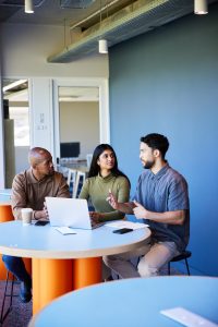 Group of diverse young businesspeople talking while working together on a laptop at a table in the lounge area of an office