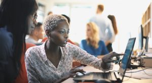 Closeup side view of mid 20's blond black woman doing her design project on a computer. She might be a software developer as well. She's sitting in front of a desktop computer and sipping a coffee while working on a computer.